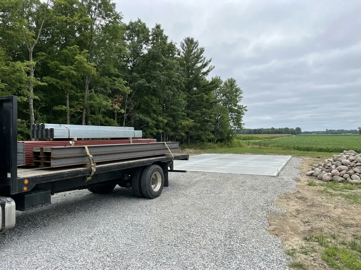 A flatbed truck delivering banded steel frame members and stacked metal panels to a building site beside a finished concrete slab.